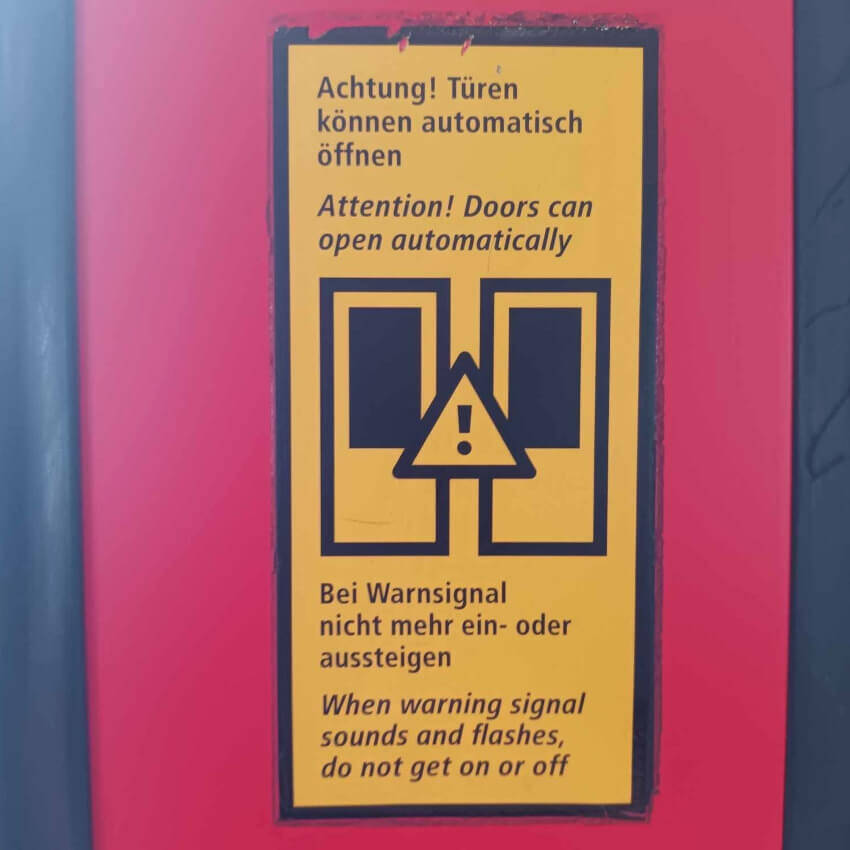 A yellow sign which shows a warning symbol over an image of the doors with text in German and English: "Attention! Doors can open automatically!"